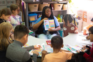 In a classroom, a high school student sits at a table with a group of elementary school student and reads "Dog Man: Big Jim Begins." The younger students follow along with their own copies.