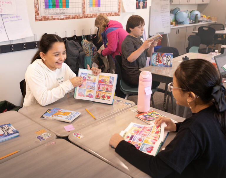 In a classroom, two students sit at a table and read their new Dog Man books together.