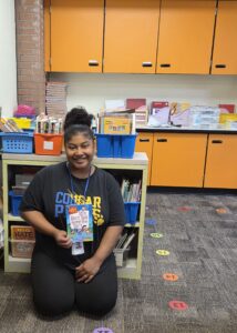 Teacher Jennifer McCoy kneels on the floor of her classroom in front of a bookshelf and holds up a book.