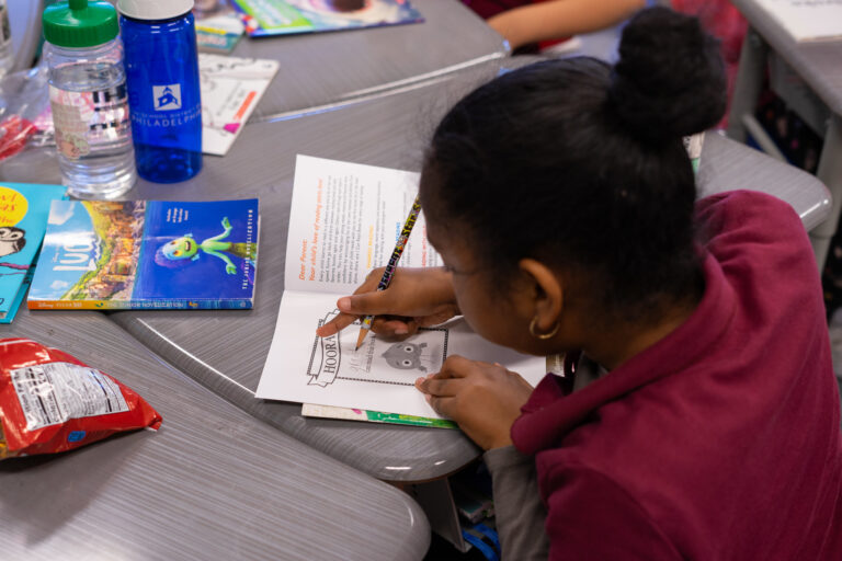 A student in a Philadelphia classroom sits at a desk and writes their name in their new Book Trust book.